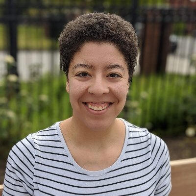 Marisa Ewing A headshot of a person with short curly brown hair, and brown skin, wearing a white striped shirt. They are smiling.