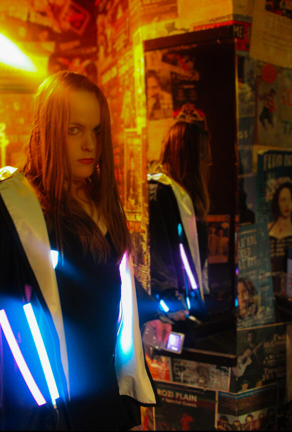 Eyre A picture of a woman in a warmly lit room. She has long brown hair, white skin, and is wearing a dark jacket, illuminated by neon lights.