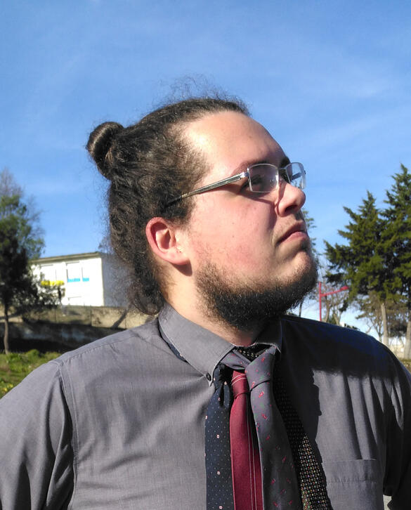 José Machado A headshot of a person with light skin and brown hair. He has a beard, a pair of glasses, and is wearing a black shirt with several ties stacked on top of each other. He is standing outsite, and staring wistfully at the sky.