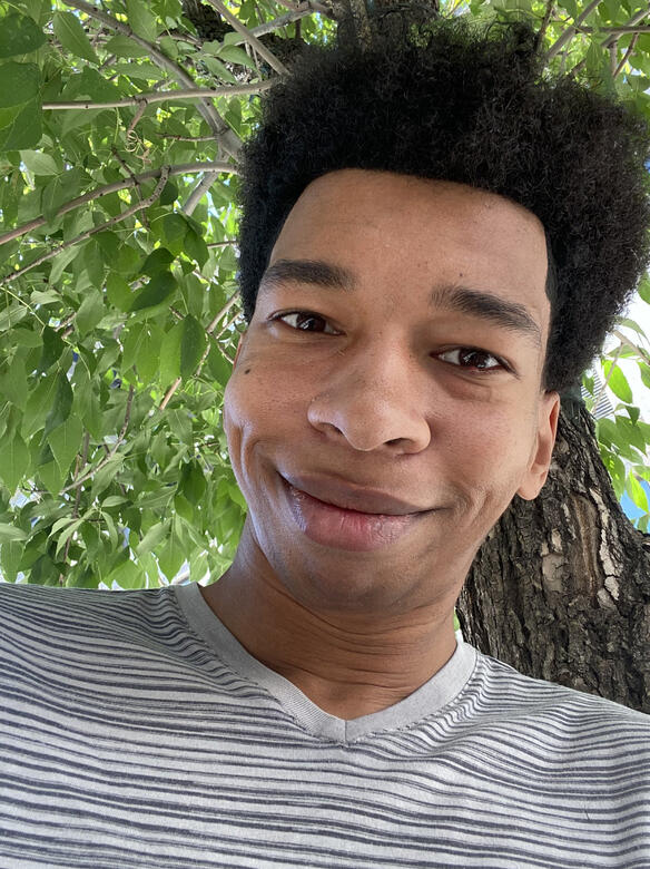 Dontae Majors A photo of a person with short curly hair and brown skin. He is wearing a white shirt with black stripes and is standing in front of a tree. He is smiling.