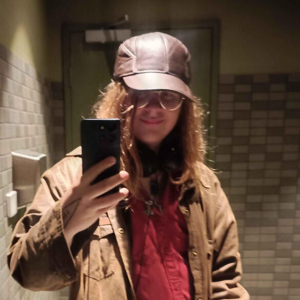 Mesabzu Wodenscild A person with light skin taking a picture in front of a mirror. They have long brown hair down to the shoulders, and they have round glasses. They are wearing a brown cap, and a brown jacket over a red shirt.