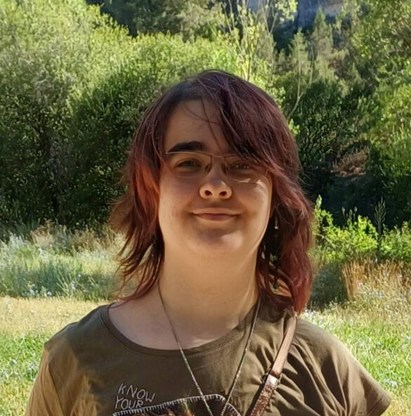 Doodles A person standing in a forest. Ey have light skin, and is wearing a dark green t-shirt. It has brownish red wavy hair, and is wearing a necklace.