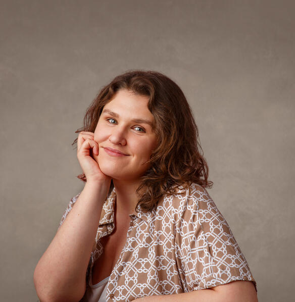 A photo of a person with light skin and brown shoulder-length hair. She is wearing a patterned light brown and white shirt. She is smiling.