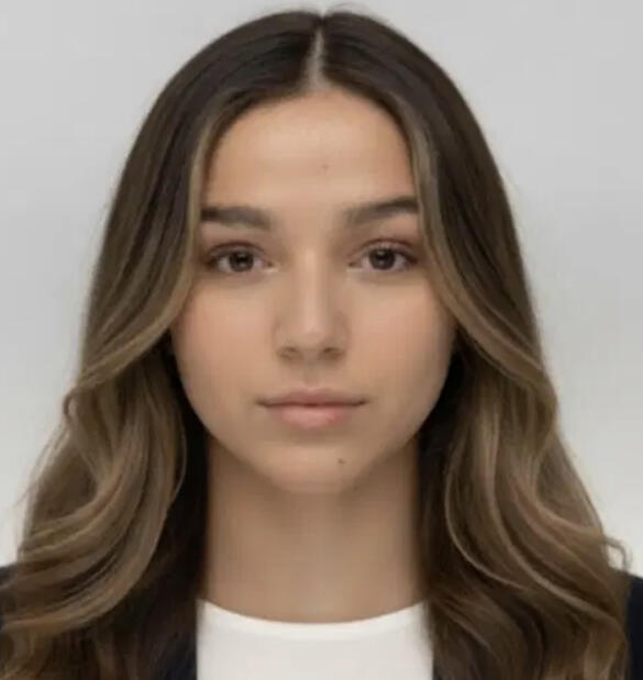 A headshot of a person with beige skin and long brown hair. She is wearing a white shirt and a black jacket.