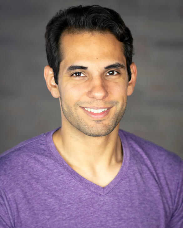 Adam Qutaishat A headshot of a person with light brown skin and short dark hair. They are wearing a purple shirt and is smiling. She is standing in front of a blurry grey backdrop.