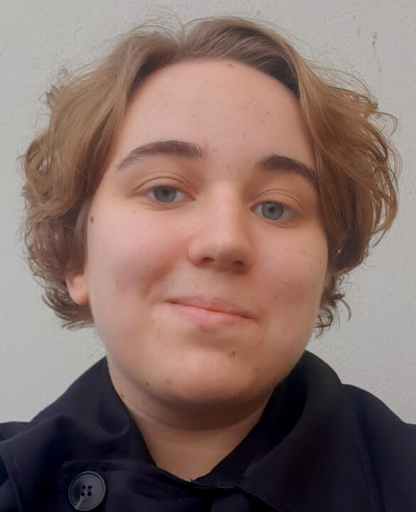 A headshot of a person with short blonde hair and light skin. They are wearing a black collared jacket and is standing in front of a white backdrop.