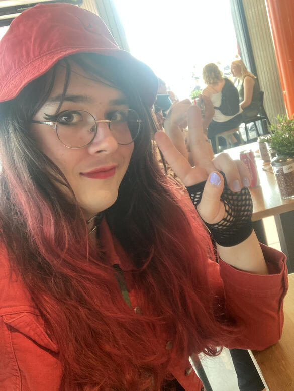 Janet G. A photo of a person doing a peace sign with her elbow on the table. She is wearing a red jacket and a red hat. She has light skin and long dark hair with the edges dyed red. She is wearing round glasses and is smiling.