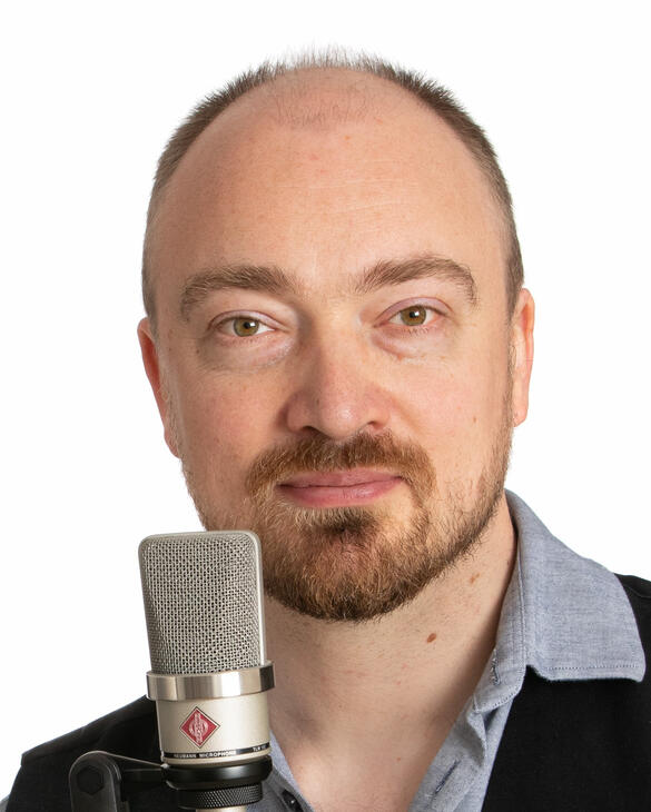A headshot of a person with light skin and a short brown beard and brown hair. He is standing in front of a white background and behind a microphone. He is smiling.