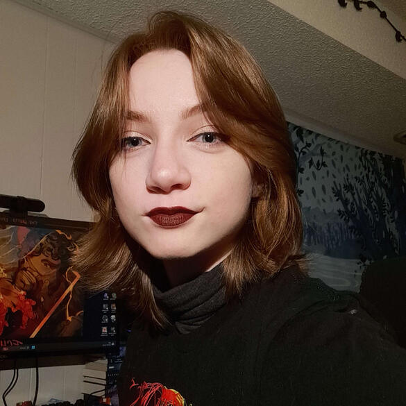 Reag Koster A photo of a person with light pale skin and chin-length brown hair. They are wearing a black outfit and is smiling.